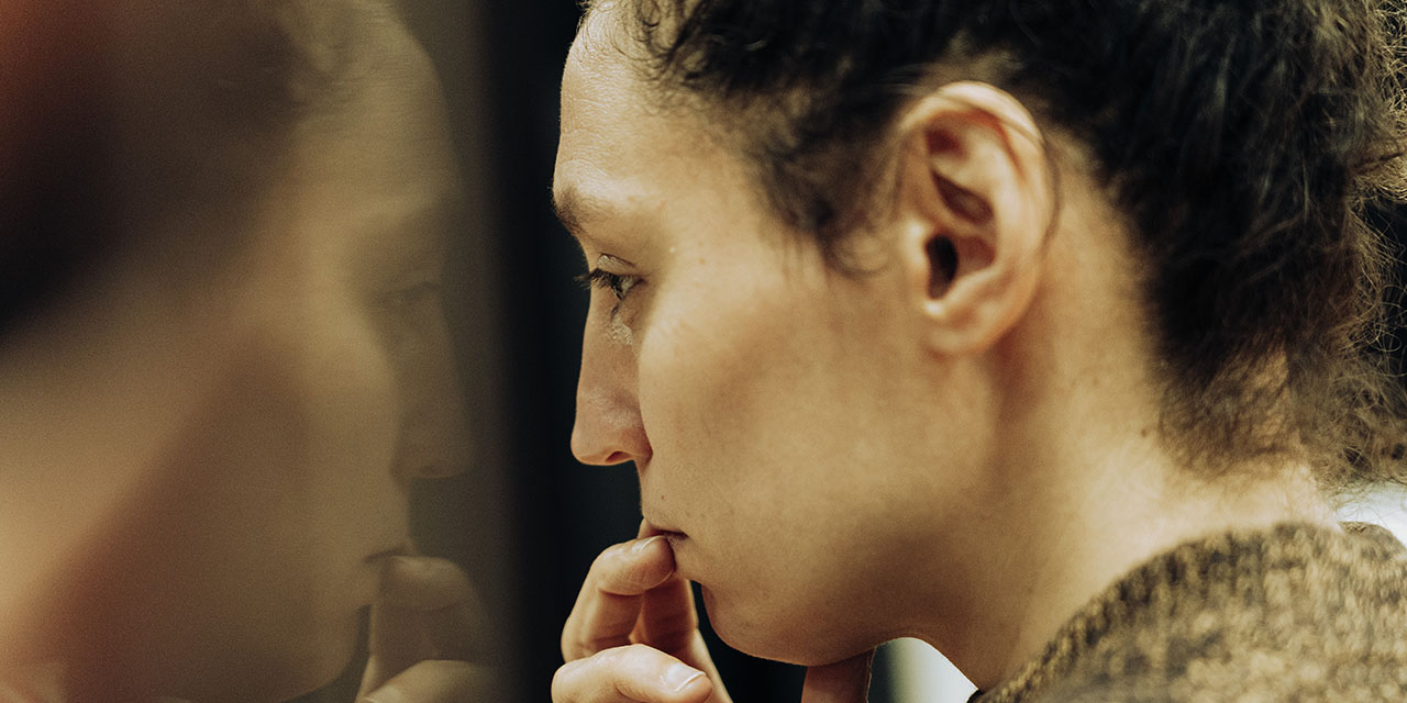Photo portrait de la comédienne, de profil elle fait face à son reflet dans un miroir légèrement tinté et à l'index posé sur sa bouche lui donnant un air songeur