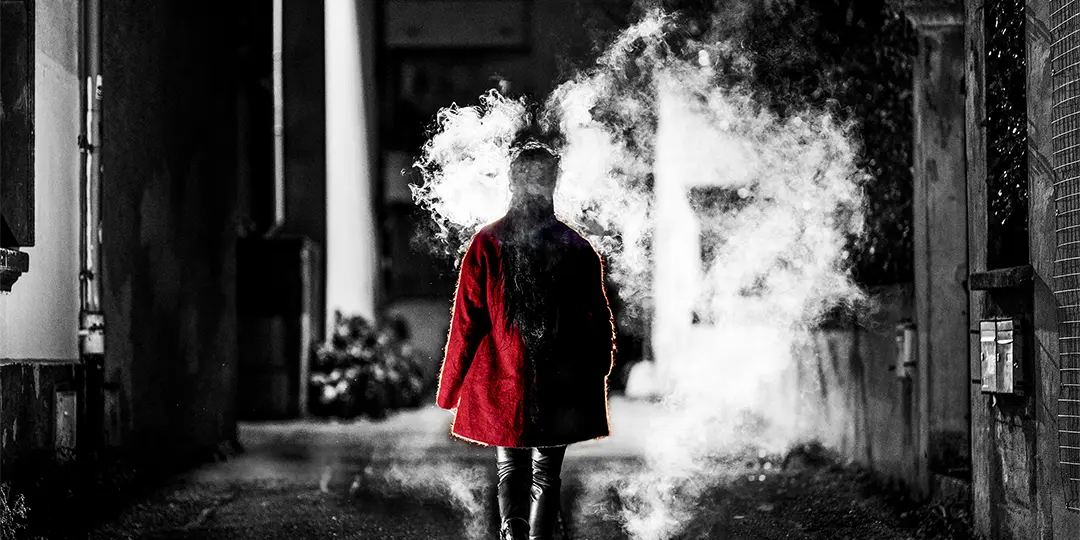 Une femme en manteau rouge marche de dos dans une ruelle sombre, entourée de fumée. 