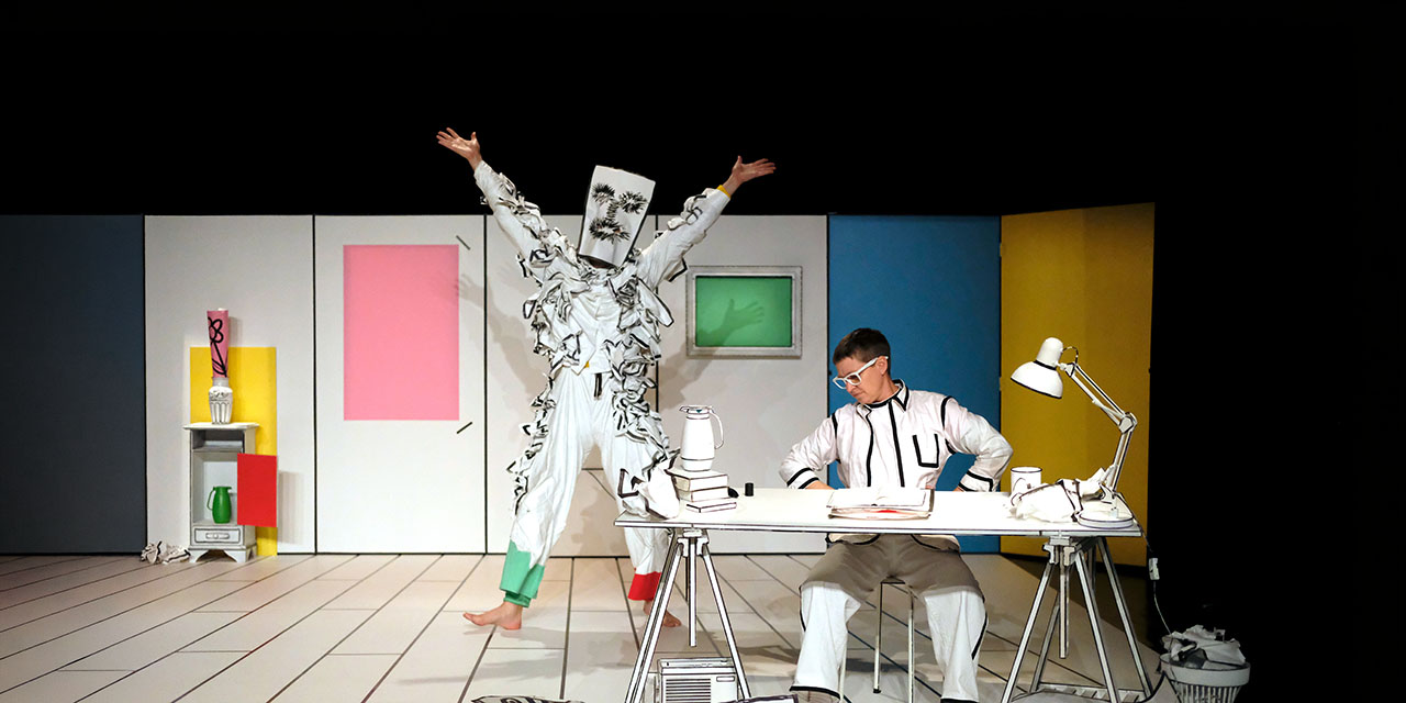 Une comédienne est assise derrière un bureau. Un danseur déguisé en bonhomme de papier danse les bras en l'air à côté d'elle.