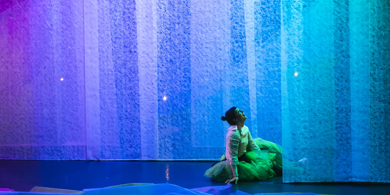 La comédienne qui interprète Blanquette est assise de profil devant des rideaux bleutés.