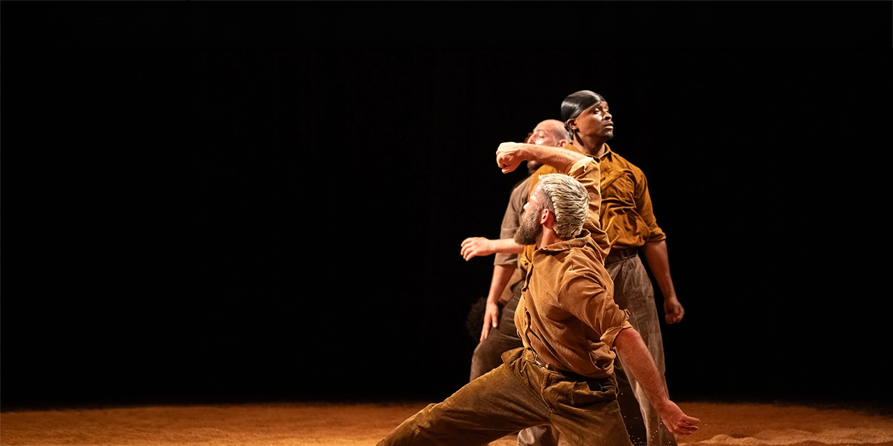 Trois danseurs sont en ligne sur un sol jonché de graines.