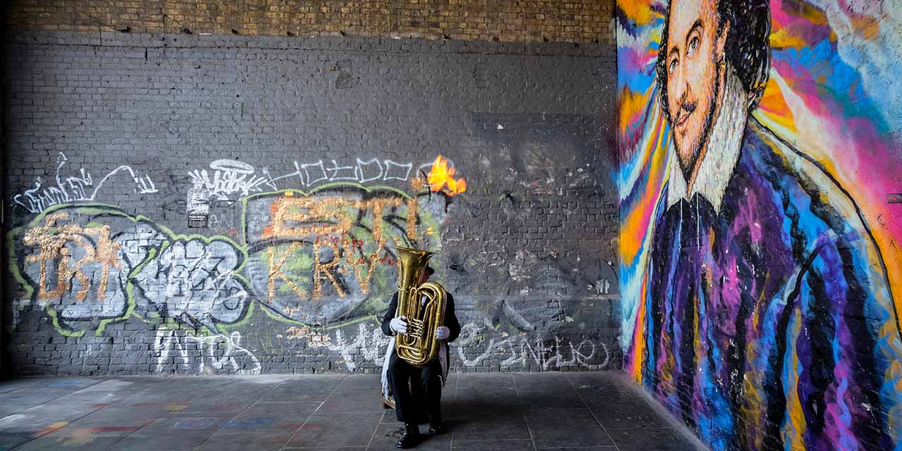 Un musicien se trouve avec son instrument devant un mur taggué où shakespeare est dessiné