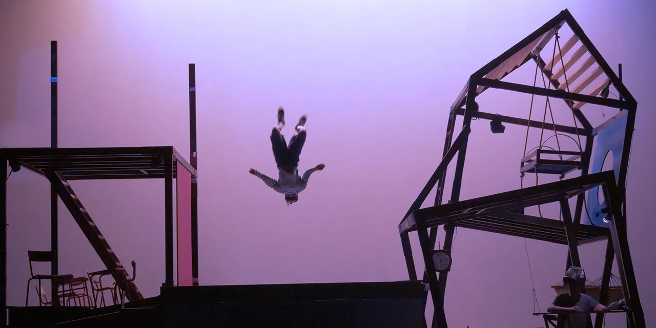 Une circassienne rebondit sur un trampoline au plateau, entouré de deux décors de maison.