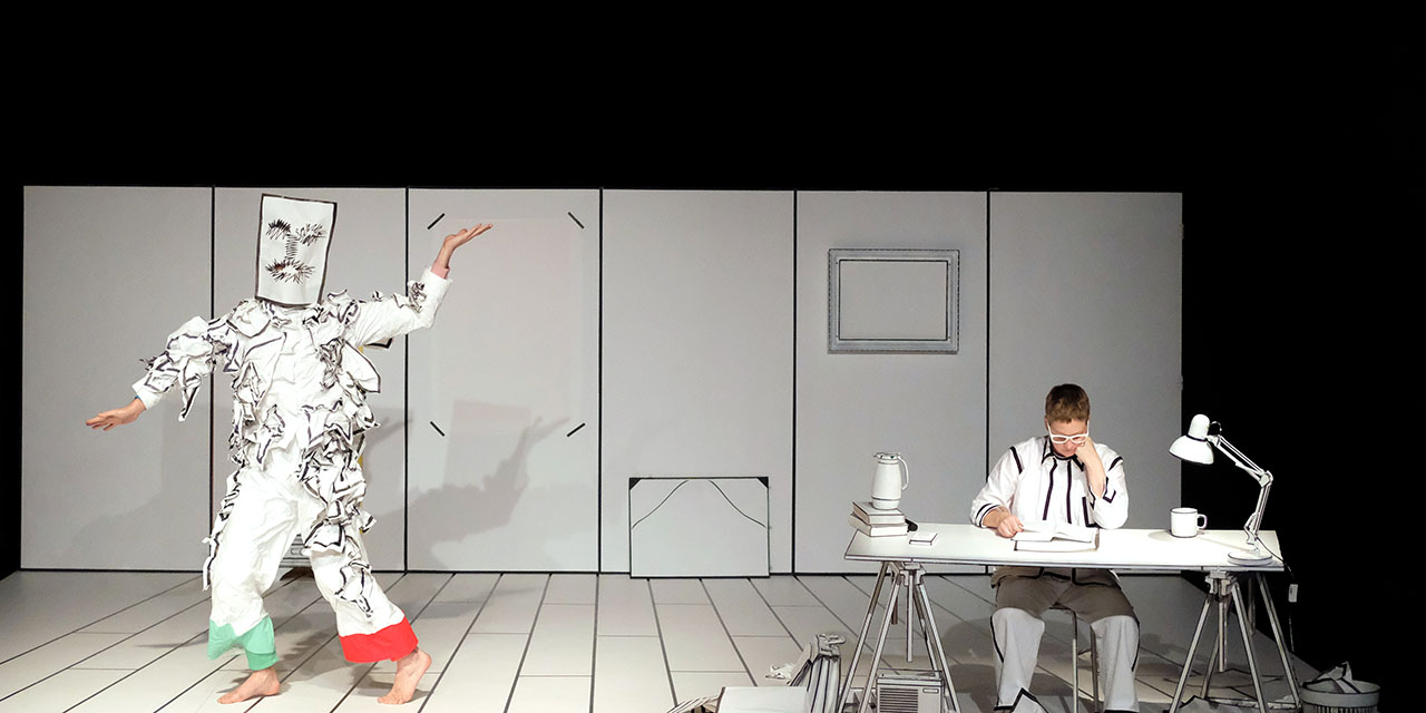 Le danseur est déguisé en bonhomme de papier dans un décor de papier, à sa droite la dessinatrice est assise en train de dessiner à un bureau