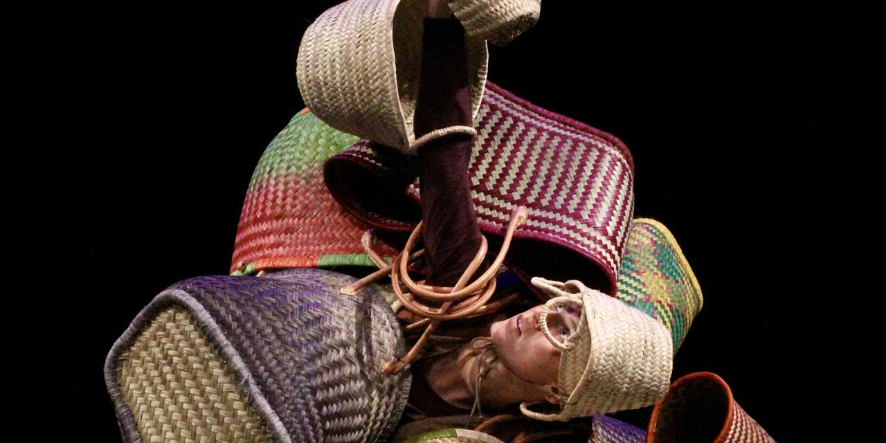 La danseuse est entourée de panier, elle en porte un comme un chapeau, d'autres autour des bras et lève son bras droit vers le haut