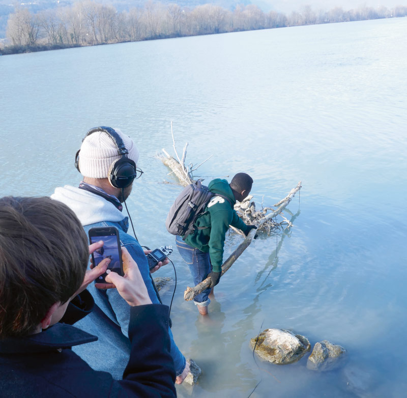 Une personne plonge un micro pendu au bout d'une branche dans l'eau tandis qu'une autre écoute au casque audio.