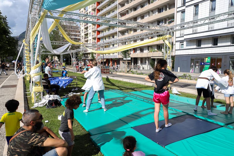 Un tatami est installé sur de la pelouse. Des personnes sont initiées au judo.