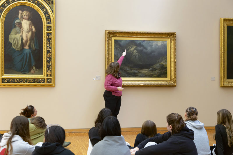 Une médiatrice donne des explications sur un tableau à un groupe de personnes assis par terre.