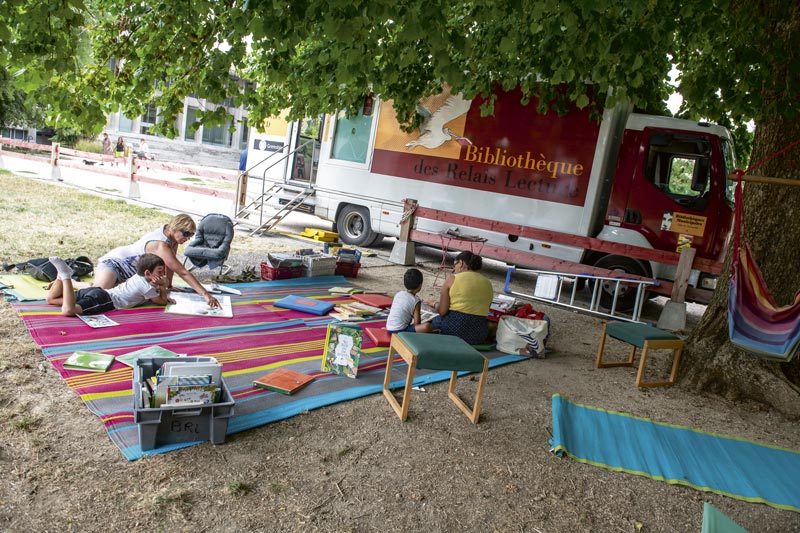 Le bibliobus est installé dans le parc Paul Mistral. Des personnes lisent sur une grande couverture.