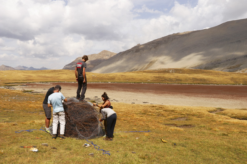 Cinq personnes préparent un sac de ferraille rouillée sur un plateau montagnard.