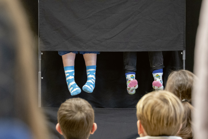 Des enfants regardent un spectacle. Sur scène, deux paires de pieds avec des chaussettes dépassent d'un rideau.