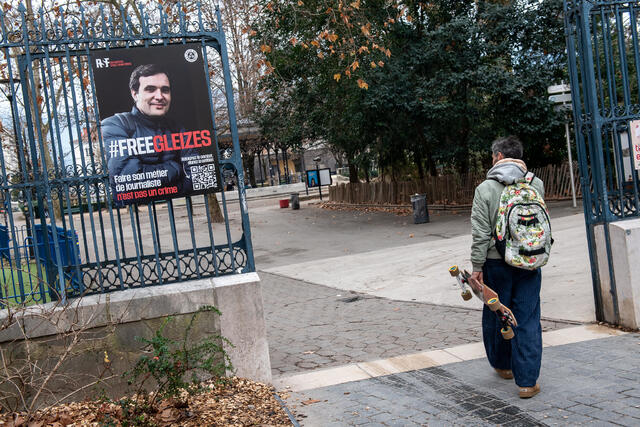 Photo d'une affiche de soutien à Christophe Gleizes, sur les grilles du jardin de ville de grenoble. Un passant regarde l'affiche en marchant.