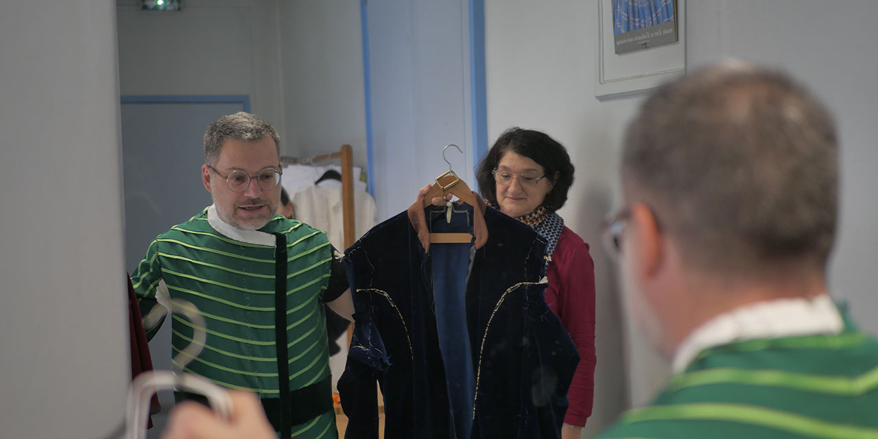 Le comédien tient deux costumes face à un miroir, à ses côtés la costumière l'observe