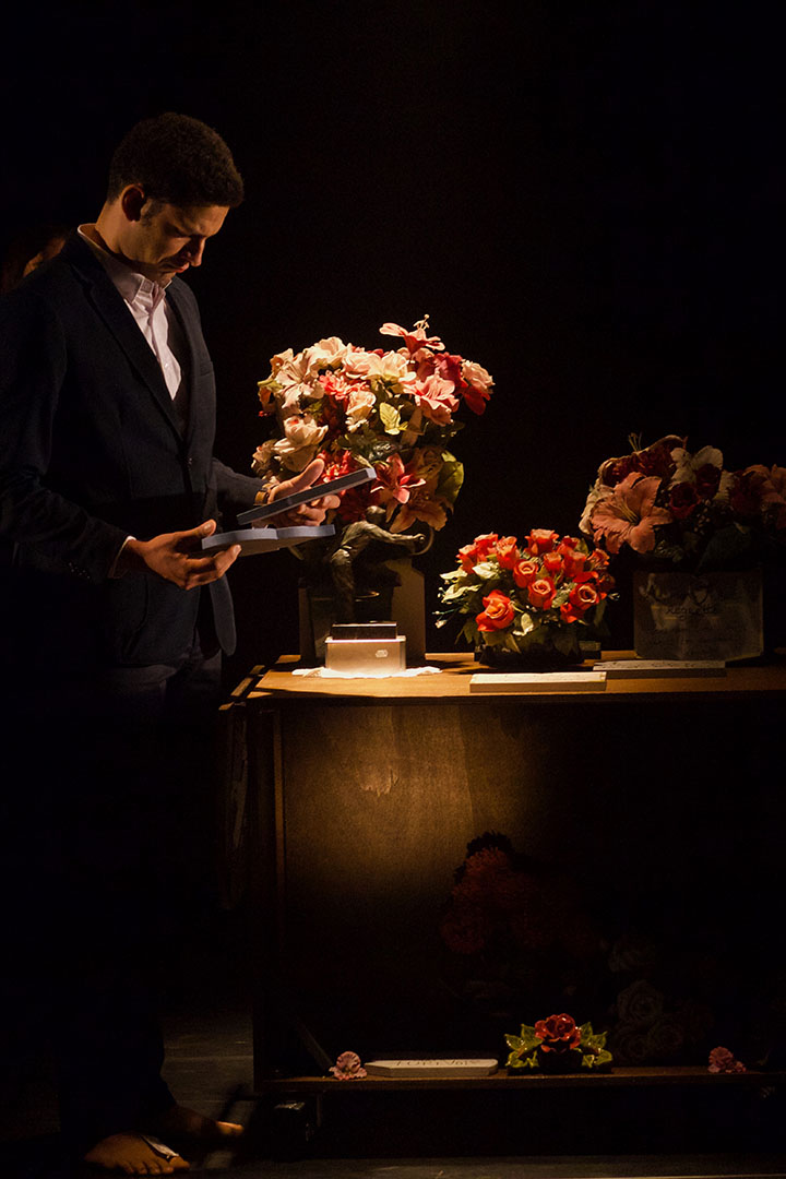 Un homme habillé en costume noir tient dans ses mains deux épitaphes. Trois bouquets de fleurs ornent un cercueil à coté de lui.
